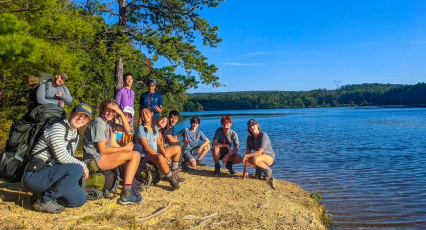 a group of teens sit next to a body of water on an outdoor expedition with outward bound